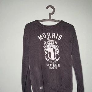 Mörkgrå stickad tröja från Morris - Snygg mörkgrå stickad tröja från Morris med stort vitt tryck och vapensköld på bröstet. Tröjan har rund halsringning, långa ärmar och ribbade muddar. Perfekt för dig som gillar klassisk och stilren design med en preppy touch.