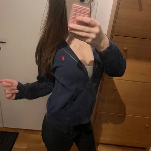 Mörkblå hoodie från Polo Ralph Lauren - Mörkblå hoodie från Polo Ralph Lauren med dragkedja framtill och klassisk röd broderad logga på bröstet. Tröjan har långa ärmar, huva och är tillverkad i mjuk bomullsmix. Perfekt för chill dagar och snygg att matcha med jeans eller leggings. helt i ny skick endast anvönd 2 gånger 🫰🏼🫰🏼 Ny pris 1200 men säljer för 1000kr🫰🏼