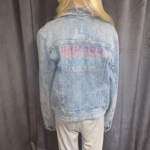 Ljusblå jeansjacka med strassdetaljer - Snygg ljusblå jeansjacka med knappar framtill och klassiska bröstfickor. På ryggen finns en cool strassdekor med texten 'UNICORN QUEEN' i rosa och blått samt en glittrig krona. Perfekt för dig som vill sticka ut med lite extra bling.