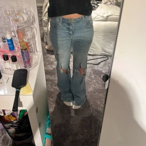Blå bootcut jeans från Zara, strl 38 - Bootcut jeans från Zara i ljusblå denim med slitna och trasiga detaljer över knäna. Klassisk femficksmodell med hög midja och raka ben som går ut lite nertill. Perfekta för en avslappnad och trendig look. Lite slitage längs ner på grund av att de är lite långa för mig.