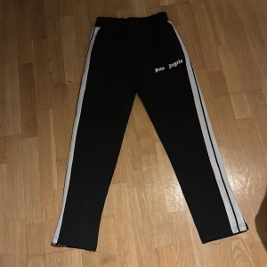 Palm Angels svarta trackpants med vita ränder - Svarta trackpants från Palm Angels med vita sidoränder och logga tryckt på vänster lår. Byxorna har elastisk midja och raka ben, tillverkade i ett mjukt och följsamt syntetmaterial. Perfekta för en streetwear-look.
