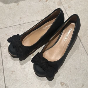 Svarta pumps med kilklack från Primavera - Snygga svarta pumps från Primavera med kilklack och dekorativ rosett framtill. Skorna har rund tå och är tillverkade i ett material som liknar skinn. Perfekta för dig som vill ha både stil och komfort med en stabil klack.