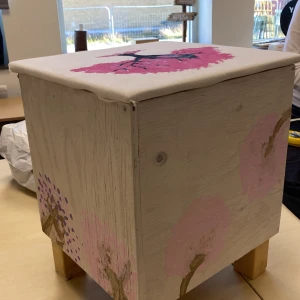 pall med förvaring - Bilderna visar en handmålad trälåda med dekorativa motiv av rosa och lila träd. Lådan har ett unikt konstnärligt utseende och står på fyra träben. Perfekt som förvaringslåda eller dekorativ möbel. Inga synliga elektroniska komponenter eller funktioner. det är en pall också