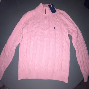 Rosa kabelstickad tröja Polo Ralph Lauren - Supersnygg rosa kabelstickad tröja från Polo Ralph Lauren med halv dragkedja i halsen och klassisk logga på bröstet. Tröjan har lång ärm, ribbade muddar och är perfekt för dig som gillar preppy stil.