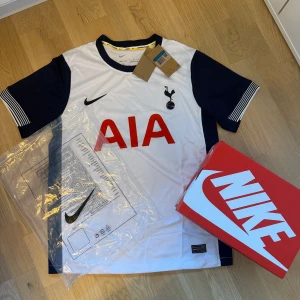 Tottenham Hotspur fotbollströja Nike fotboll jersey - Snygg Tottenham Hotspur matchtröja från Nike med korta ärmar. Tröjan är vit med marinblå detaljer på ärmar och krage, samt röd AIA-logga på bröstet. Materialet är lätt och ventilerande polyester, perfekt för träning eller match. Ikoniskt klubbmärke på bröstet.