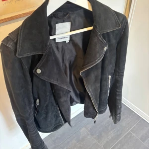 Svart mockajacka från J.Lindeberg - Snygg svart mockajacka från J.Lindeberg med klassisk bikerstil. Jackan har breda slag, silverfärgade dragkedjor och knappar samt långa ärmar med dragkedjor vid ärmslut. Materialet är mocka och jackan har en cool, tidlös look. Hål innanför armhålan som går att sy ihop. inget man märker/ ser 