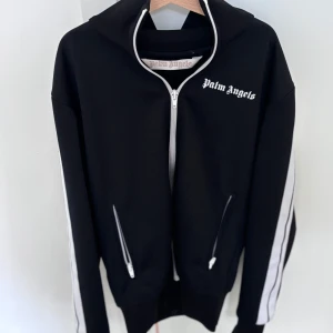 Palm Angels svart hoodie med dragkedja - Svart hoodie från Palm Angels med vit dragkedja och vita ränder längs ärmarna. Klassisk logga på bröstet och två fickor med dragkedja. Str XS äkta såklart