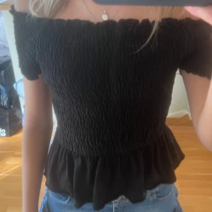 Svart offshoulder topp med volang - Säljer en svart offshoulder topp med smockad överdel och volang nertill. Toppen är tight upptill och har en snygg rynkad struktur. Perfekt till jeans eller kjol för en trendig look.