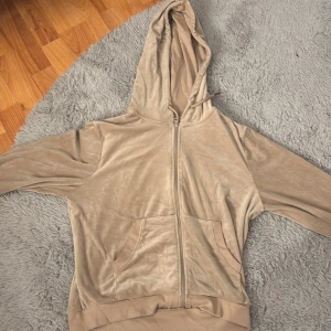 Beige velour hoodie med dragkedja - Mjuk och skön beige hoodie i velour med dragkedja framtill och huva med snörning. Tröjan har två stora fickor framtill och ribbade muddar vid ärmslut och nederkant. Perfekt för en chill och avslappnad stil.
