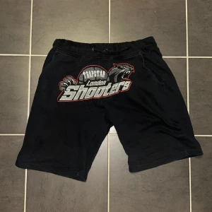Trapstar London Shooters shorts svart - Svarta shorts från Trapstar med stort tryck framtill där det står 'London Shooters' och en panter. Modellen har snörning i midjan och är tillverkad i mjuk bomull, perfekt för en sportig och avslappnad stil.
