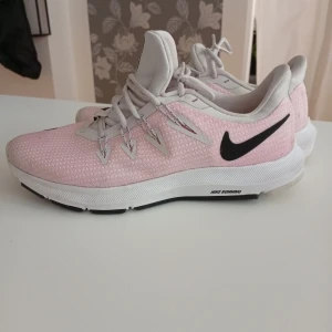 Nike rosa och vita löparskor - Säljer ett par snygga Nike löparskor i ljusrosa och vitt med svart swoosh-logga. Skorna har mesh-ovandel för bra andningsförmåga, snörning och en vit sula med greppvänligt mönster. Perfekta för dig som gillar att springa eller vill ha sportiga sneakers.