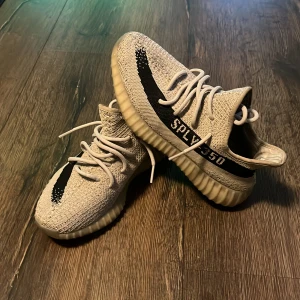 Adidas Yeezy Boost 350 V2 Zebra - Säljer ett par Adidas Yeezy Boost 350 V2 Zebra med stickad ovandel i beige och svart, klassisk SPLY-350 text på sidan och vit snörning. Skorna har en tjock, ribbad sula och är riktigt bekväma med sin boost-dämpning. Perfekta för dig som vill sticka ut med en ikonisk sneaker.