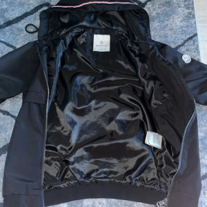 Svart vindjacka från Moncler med huva - Svart vindjacka från Moncler med glansig insida, huva med rödvit rand och dragkedja framtill. Jackan har långa ärmar, två fickor med dragkedja och Moncler-logga på ärmen. Perfekt för dig som vill ha en stilren och sportig look.