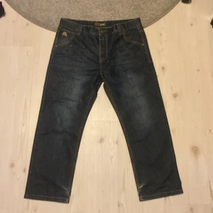 Mörkblåa jeans från Jeep med brodyr - Snygga mörkblå jeans från Jeep med gul broderad logga på fickan och bakficka. Klassisk femficksmodell med kontrastsömmar och läderpatch bak i midjan. Raka ben och normal passform, perfekta till vardagslooken.