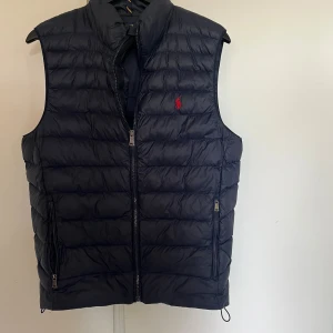 Mörkblå Polo Ralph Lauren väst - Snygg blå dunväst från Polo Ralph Lauren med quiltad design och dragkedja framtill. Västen har två fickor med dragkedja och klassisk röd logga broderad på bröstet. Perfekt för lager-på-lager och stilren look.