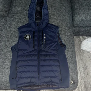 Marinblå dunväst från Sail Racing - Snygg marinblå dunväst från Sail Racing med huva och dragkedja framtill. Västen har broderad logga på bröstet, patch med 'Antarctica Expedition' och två sidofickor med dragkedja. Perfekt för lager-på-lager och sportig stil.