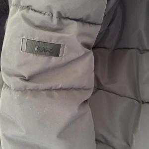 Grå pufferjacka från Michael Kors L - Snygg grå pufferjacka från Michael Kors med huva och dragkedja framtill. Jackan har vattenavvisande material och isolering som håller dig varm. Stora fickor och diskret MK-logga på ärmen ger en clean look. Perfekt för kalla dagar när du vill vara både varm och stilren.