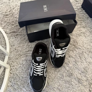 Svarta Dior sneakers med vit sula - Säljer ett par svarta Dior sneakers med vit och grå detalj samt tjock vit sula. Skorna har tydliga Dior-loggor på plösen och sidan, samt vita snören. Snygg och modern design som sticker ut, perfekt för dig som gillar exklusiva märken och streetstyle.
