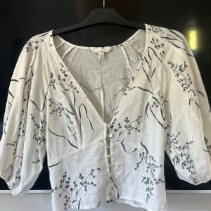 Vit blommig blus från H&M i linneblandning - Superfin vit blus från H&M i linneblandning med svarta broderade blommor och växtmönster. Blusen har v-ringning, knappar framtill och puffiga trekvartsärmar. Perfekt för dig som gillar en luftig och romantisk stil.