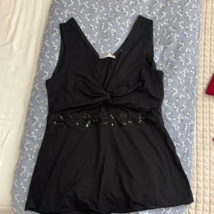 Svart linnetopp med paljettdetalj - Svart ärmlös linnetopp med djup v-ringning både fram och bak. Toppen har en snygg twistad detalj framtill och ett brett band med svarta paljetter och broderier i midjan. Materialet känns mjukt och stretchigt, perfekt för att lyfta en outfit.