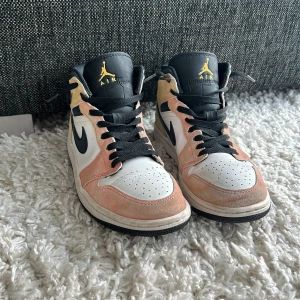 Nike Air Jordan 1 Mid - Nike Air Jordan 1 Mid sneakers med vit läderbas, pastellrosa och gul mocka, svarta detaljer och snörning. Klassisk svart  AirJordan-logga på sidan. Hög modell med gul Jordan logga. 