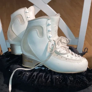 Vita konståkningsskor Edea 240 - Säljer ett par vita konståkningsskor från Edea i storlek 240. Skorna har snörning, gulddetaljer vid hälen och är tillverkade i syntetmaterial med meshpartier. Klassisk design med rund tå och vadderad insida för extra komfort. Säljer dessa pga bytte till piano. Jätte fina och bra skick! Skenan är jätte bra och fungerar bra till dubbel hopp. Kom med pris förslag!