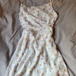Vit blommig omlottklänning från H&M - Supersöt vit kortklänning från H&M med små blommor i rosa, orange och gult. Klänningen har smala axelband och knytning i ryggen. Perfekt för sommarens alla tillfällen!