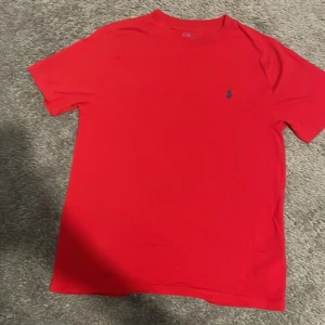 Röd t-shirt från Polo Ralph Lauren - Röd t-shirt från Polo Ralph Lauren med klassisk broderad logga på bröstet. Enkel och stilren design med rund hals och korta ärmar. Tillverkad i mjuk bomull som känns skön mot huden. Perfekt för dig som gillar ikoniska märken och clean look.