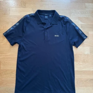 Svart pikétröja från BOSS - Snygg svart pikétröja från BOSS med klassisk krage och knäppning framtill. Tröjan har korta ärmar och diskreta BOSS-loggor längs axlarna. Perfekt för en stilren och sportig look.