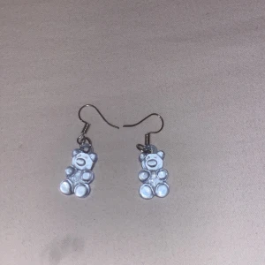 Söta örhängen med nallebjörnar - Unika örhängen med små vita nallebjörnar i plast. Perfekta för dig som vill ha en lekfull och gullig detalj till din stil. Krokar i metall och nallarna har en glansig finish som fångar ljuset snyggt.