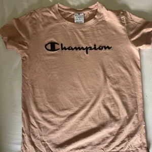 Beige Champion t-shirt XS - Snygg beige t-shirt från Champion i storlek XS. Klassisk modell med rund halsringning och svart Champion-logga tryckt framtill. Tillverkad i mjuk bomull som är skön mot huden. Perfekt för en avslappnad och sportig stil.