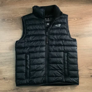 Hollister Väst - Svart quiltad dunväst från Hollister Puffer Collection med återvunnet fyllnadsmaterial. Västen har ståkrage, två fickor med dragkedja och ett broderat Hollister-logo på bröstet. Snyggt horisontellt mönster och dragkedja framtill.