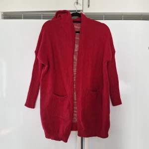 Röd stickad kofta med huva och text stl 38 - Mysig röd stickad kofta med huva och två stora fickor framtill. På ryggen finns ett beige mönster med texten 'LIVE SIMPLY' och hjärtan. Långa ärmar med ribbade muddar och avslappnad passform. Perfekt för lager-på-lager under kyliga dagar. Aldrig använd. 