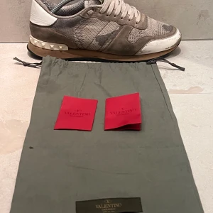 Valentino Garavani Rockrunner sneakers - Grå och vita Valentino Garavani Rockrunner sneakers med coolt kamouflagemönster, detaljer i mocka och mesh samt ikoniska nitar på hälen. Skorna har snörning och rund tå, samt en kontrasterande sula i beige och svart. Perfekta för dig som gillar streetstyle med lyxig touch.