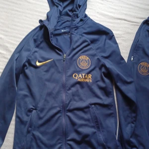 PSG träningsset Nike blå/gul - Säljer ett Paris Saint-Germain träningsset från Nike i marinblått med gula detaljer. Setet består av en huvtröja med dragkedja och byxor, båda med PSG-logga och Qatar Airways-tryck. Materialet är lätt och andas, perfekt för fotbollsträning. Barn storlek 128-137 . 8-10 år .