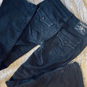 Svarta True Religion Becca Bootcut Jeans - Svarta jeans från True Religion, modell Becca Mid Rise Bootcut. Klassisk femficksdesign med snygga detaljerade bakfickor med lock och knappar. Köpta för 1300kr och knappt använt därav priset, dom ser fortfarande nya ut och dom är inte i närheten av trasiga nån stans.