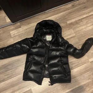 Svart Moncler pufferjacka med huva - Säljer en svart pufferjacka från Moncler med glansig finish och huva. Jackan har dragkedja, stora fickor med lock och Moncler-logga på ärmen. Insidan har ett unikt serietryck. Perfekt för kalla dagar och riktigt snygg streetstil.