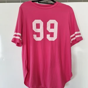 Rosa t-shirt med tryck 99 L - Cool rosa t-shirt med vita ränder på ärmarna och stort tryck med siffran 99 på bröstet. Oversized passform och sportig vibe, perfekt för en avslappnad look. Gjord i mjukt bomullstyg.. Använd 1 gång. 