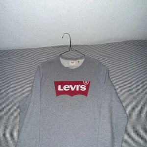 Grå Levi's sweatshirt med rött tryck  - Säljer en grå sweatshirt från Levi's i storlek XS. Tröjan har ett stort rött och vitt Levi's-logotyptryck på bröstet, rund halsringning och långa ärmar. Tillverkad i mjuk bomullsmix, perfekt för en chill och stilren look.