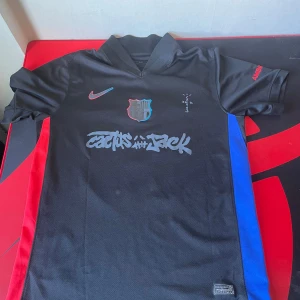 FC Barcelona Cactus Jack Lamine Yamal tröja - Säljer en svart FC Barcelona x Cactus Jack fotbollströja från Nike med Lamine Yamal och nummer 19 på ryggen. Tröjan har röda och blå sidopaneler, klubbmärke på bröstet och Cactus Jack-tryck framtill. Tillverkad i ventilerande polyester med korta ärmar.