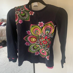Barn Desigual långärmad blommig tröja - Supersnygg långärmad tröja från Desigual med färgglada blommiga mönster framtill. Basen är mörkblå och tröjan har coola detaljer som rosa spetskant nertill och små rosa inslag vid ärmsluten. Perfekt för dig som gillar unika och färgstarka plagg!