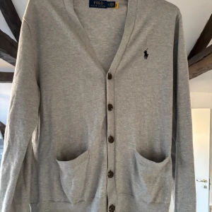 Grå kofta från Polo Ralph Lauren - Snygg grå kofta från Polo Ralph Lauren med klassisk v-ringning och knappar framtill. Tröjan har två fickor och den ikoniska loggan broderad på bröstet. Perfekt att slänga över en t-shirt för en chill look.