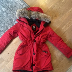 Röd Canada Goose parkas med päls - Snygg röd parkas från Canada Goose med pälskantad huva och klassisk logga på ärmen. Perfekt för kalla dagar med sin varma dunfyllning och justerbar midja för schysst passform. Lite skitig på ärmar men går bort i tvätt, Liten i strl