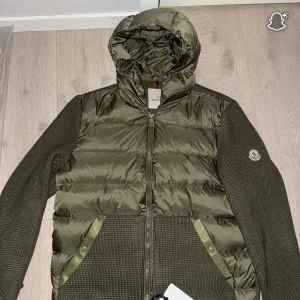 Moncler cardigan Helt ny  - Säljer en olivgrön pufferjacka från Moncler med stickade ärmar och huva. Jackan har quiltad framsida i glansigt material, två fickor med tryckknappar och dragkedja framtill. Moncler-logga på ena ärmen. Perfekt för höst och vinter! Säljer min moncler cardigan 2025 modellen. Anledningen är att den är för stor. De är en size 4 så storlek 4, stl L. Kvaliteten är helt galen. Jackan är en Akp