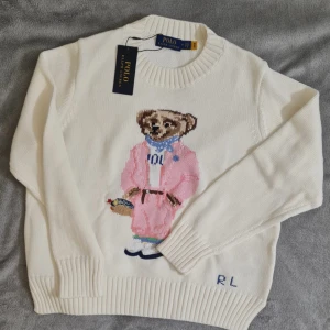 Vit stickad tröja Ralph L - Vit stickad tröja från Ralph L med ikonisk björn på bröstet, klädd i rosa kappa och blå scarf. Tröjan har rund halsringning, ribbade muddar och RL-broderi nertill. Perfekt statement-piece för dig som gillar preppy stil. Unisex storlek S 