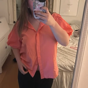 Korallrosa kortärmad skjorta från marta - Säljer en korallrosa skjorta från marta med korta, puffiga ärmar och knappar framtill. Skjortan har en lös passform och är tillverkad i ett lätt bomullstyg, perfekt för varmare dagar. Snygg krage och enkel att styla till olika outfits.