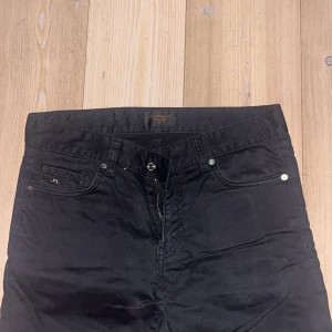 Svarta jeans från J.Lindeberg - Svarta jeans från J.Lindeberg med klassisk femficksdesign och diskret logga på myntfickan. Byxorna har normal passform och är tillverkade i ett mjukt bomullsmaterial med lite stretch. Perfekta för dig som gillar stilrena och enkla jeans.