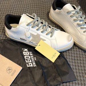 GGDB vita sneakers med grå stjärna - Snygga vita sneakers från GGDB med grå snörning och en ikonisk grå stjärna på sidan. Skorna har svart hälparti med GGDB-tryck och en platt sula i beige och svart. Tillverkade i skinn med detaljer i mocka. Perfekt för dig som gillar stilrena och coola sneakers.