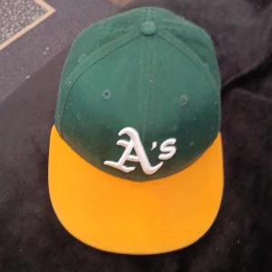 Oakland Athletics keps från New Era - Snygg keps från New Era med Oakland Athletics A's-logga framtill. Kepsen har en mörkgrön krona och gul böjd skärm, klassisk basebollstil. Broderad logga på sidan och ventilerande hål upptill. Perfekt för dig som gillar sportig streetwear.