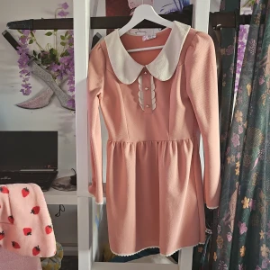 Söt rosa klänning med vit krage - Supersöt kort rosa klänning från Seifini med långärmade ärmar och stor vit krage. Klänningen har dekorativa pärlknappar framtill och scallop-detaljer vid knäppningen. Kjolen är lätt utställd och har vit spetskant längst ner. Perfekt för dig som gillar gullig och unik stil!